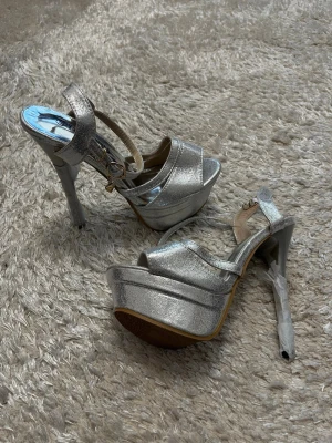 Silvriga platåpumps med höga klackar - Säljer ett par oanvända silvriga pumps med platå och höga stilettklackar. Skorna har öppen tå och justerbar rem med spänne runt ankeln. Materialet är glansigt och metallic, perfekt för att sticka ut. Yttersulan är ljusbrun och insidan har metallicfinish.