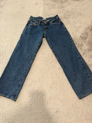Blå raka jeans med hög midja - Säljer ett par klassiska blå jeans med rak passform och hög midja. Jeansen har fem fickor, bälteshällor och stängs med dragkedja och knapp. Materialet är kraftig denim i bomull. Perfekta för en avslappnad och trendig look.
