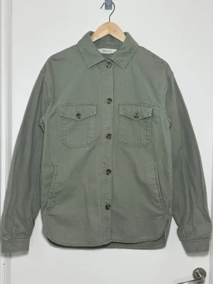 Grön skjortjacka från H&M - Grön skjortjacka från H&M med klassisk krage, två bröstfickor med lock och knappar samt raka ärmar med knapp vid ärmslut. Tillverkad i ett kraftigare bomullstyg som ger en avslappnad och stilren look. Perfekt att bära öppen eller stängd.