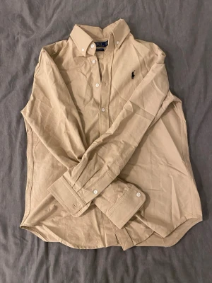 Beige skjorta från Polo Ralph Lauren - Klassisk beige skjorta från Polo Ralph Lauren i custom slim fit. Skjortan har button-down krage, svarta broderade loggan på bröstet och vita knappar. Tillverkad i mjuk bomull och passar perfekt till en avslappnad men ändå stilren look. Skjortan är använd en gång och i nyskick! Seriösa bud kan godkännas.