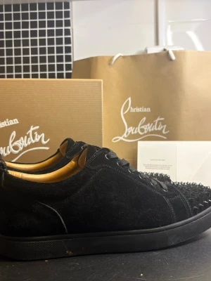 Louboutin skor - Säljer ett par svarta sneakers från Christian Louboutin i mocka med svarta nitar på tån. Klassisk låg modell med svarta skosnören och diskret logga på insidan. Snygg och edgy design som sticker ut.