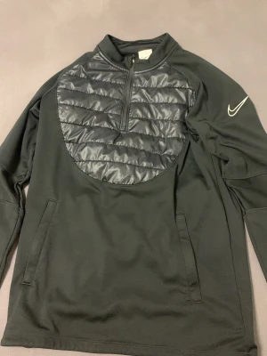 Svart Nike therma tröja med quiltad detalj - Snygg svart långärmad tröja från Nike therma med quiltad, vadderad panel över bröstet och halv dragkedja. Tröjan har fickor på sidorna och en vit Nike-logga på ärmen. Perfekt för dig som gillar sportig och modern stil.