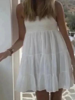 Vit klänning - Söt vit kortklänning med volanger och smock upptill. Klänningen är ärmlös och har en luftig passform, perfekt för varma dagar. Tillverkad i ett lätt material som ger en avslappnad och trendig look.