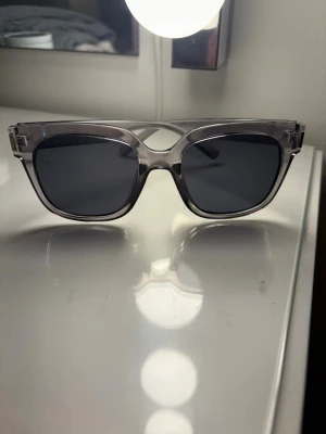 Grå transparenta solglasögon - Snygga solglasögon med transparent grå båge och mörka glas. Rektangulär form som ger en modern vibe och passar till de flesta outfits. Bågarna är i plast och har en stilren design utan synliga loggor eller detaljer.