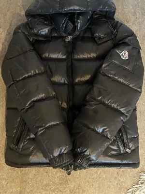 Svart dunjacka från Moncler med huva - Snygg svart dunjacka från Moncler med glansig finish och stor huva. Jackan har dragkedja framtill, två sidofickor med dragkedja och Moncler-logga på ärmen. Puffig modell som håller dig varm under kalla dagar. Perfekt för dig som vill ha både stil och funktion.