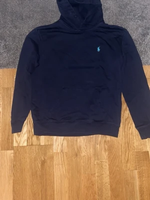 Mörkblå hoodie från Polo Ralph Lauren - Mörkblå hoodie från Polo Ralph Lauren med klassisk huva och känguruficka framtill. Liten turkos logga broderad på bröstet. Tillverkad i mjukt bomullsmaterial och har ribbade muddar vid ärmslut och nederkant. Perfekt för en avslappnad och stilren look. Storlek L men har blivit liten på fel tvätt så den är en storlek S