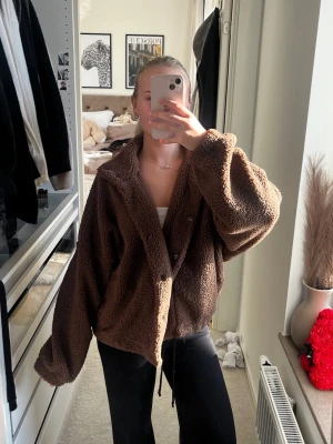 Brun oversized teddyjacka - Mysig och oversized brun teddyjacka med dragkedja och knappar framtill. Jackan har en hög krage och är tillverkad i fluffigt teddy-material som håller dig varm. Perfekt för att skapa en chill och trendig look under kalla dagar.