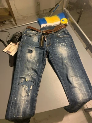 Blå slitna jeans med färgstänk - Säljer ett par blå dsquared2 jeans med slitningar, hål och coola färgstänk för en edgy streetlook., normal midja och dragkedja. Perfekta för dig som gillar en unik och trendig vibe.  ””Priset e inte fast så går att pruta