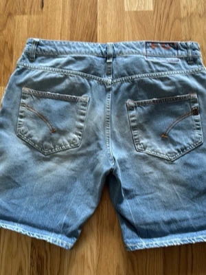 Blå jeansshorts med bakfickor från Dondup  - Snygga blå jeansshorts från Dondup med klassisk femficksdesign och coola detaljer på bakfickorna. Shortsens denim är ljusblått och har kontrastsömmar. Perfekta för varma dagar och enkel att matcha med t-shirt eller hoodie. Se bilder då ena öglan är trasig men en skräddare har lagat det.