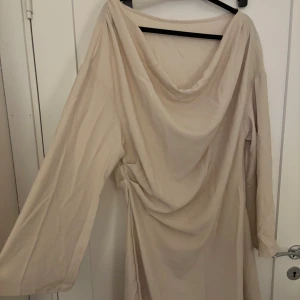 Beige långklänning med drapering - Elegant beige långklänning med draperad halsringning och långa ärmar. Klänningen har en mjuk och flytande passform och är tillverkad i ett lätt material som ger ett snyggt fall. Perfekt för dig som gillar stilrena och sofistikerade looks.