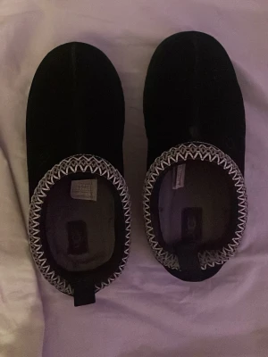 Svarta tofflor med mönstrad kant - Mysiga svarta tofflor från Decathlon med vit och grå zigzag-mönstrad kant runt öppningen. Tofflorna har en mjuk ovandel och en räfflad sula för bättre grepp. Perfekta för att hålla fötterna varma hemma. Kan även användas på sommaren. 