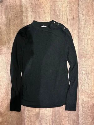 Svart ribbad långärmad topp MNG - Svart ribbad långärmad topp från MNG med dekorativa pärlknappar på axeln. Hög hals och figurnära passform, perfekt att styla med jeans eller kjol. Enkel men snygg detalj med knapparna på axeln.