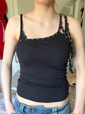 Svart ribbad linnetopp med nitar - Trendig svart ribbad linnetopp från Bershka med asymmetrisk design och coola nitar längs kanten. Ena axelbandet har extra detaljer med metallringar och snörning, samt en liten spetsdetalj vid urringningen. Perfekt för en edgy look.