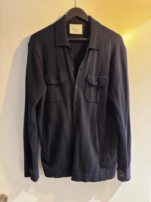 Maglia Studios Kofta - Stilren marinblå kofta från Maglia Studios med knäppning framtill och två bröstfickor med lock. Koftan har klassisk krage och långa ärmar med knapp vid ärmslut. Perfekt för lager-på-lager och enkel att matcha. Mjuk och bekväm känsla.