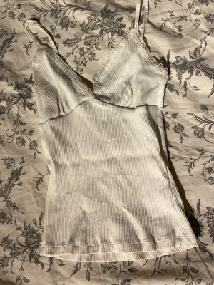 Vit ribbad linnetopp med spets - Snygg vit ribbad linnetopp med smala axelband och spetsdetaljer vid bysten. Toppen har en figurnära passform och är perfekt att styla till jeans eller kjol. Enkel men ändå söt design som passar till många olika looks.