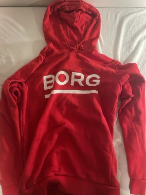 Röd hoodie från Björn Borg - Snygg röd hoodie från Björn Borg med stor vit logga på bröstet. Modellen heter Stanley Hood och har klassisk huva med dragsko samt ribbade muddar. Perfekt för en avslappnad streetstyle-look. I väldigt fint skick varsamt använd. Inga fläckar och inte sliten heller.