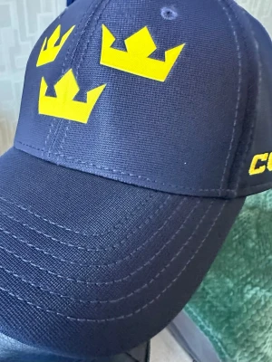 Mörkblå Tre Kronor keps - Snygg mörkblå keps från CCM med Tre Kronor-loggan i gult framtill. Kepsen har böjd skärm, broderade detaljer och justerbar passform. Perfekt för dig som gillar hockey och vill visa ditt stöd för Sverige.