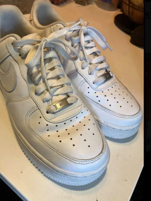 Nike Air Force 1 vita sneakers - Klassiska Nike Air Force 1 sneakers i helvitt läder med perforerad tå, vit sula och matchande vita skosnören. Ikonisk Nike Swoosh på sidorna och metallbricka med 'AF1' på snörningen. Perfekt för en clean och stilren look.