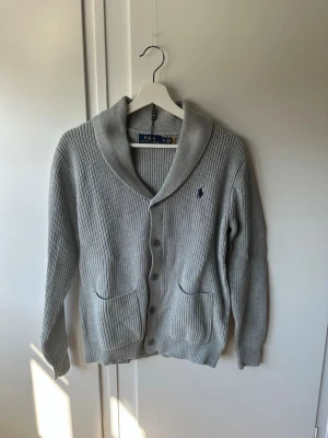 Grå kofta från Polo Ralph Lauren - Snygg grå kofta från Polo Ralph Lauren med sjalkrage, knäppning framtill och två fickor. Klassisk ribbstickad design och broderad logga på bröstet. Perfekt för lager-på-lager och chill stil.