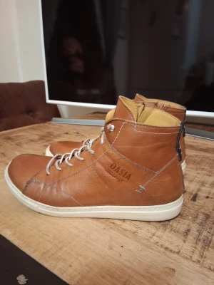 Bruna höga sneakers från Dasia - Snygga höga sneakers i brunt läder från Dasia med vita sulor och vita snören. Skorna har en stilren design med svarta detaljer baktill och dragkedja på sidan för enkel påtagning. Perfekta för dig som vill ha en clean och trendig look.