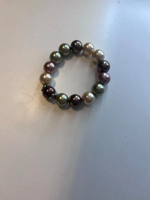 Flerfärgat pärlarmband - Säljer ett snyggt armband med stora pärlor i olika färger som grön, lila, brun och krämvit. Pärlorna är runda och sitter på en elastisk tråd, vilket gör det enkelt att ta på och av. Perfekt accessoar för att ge din outfit lite extra färg.