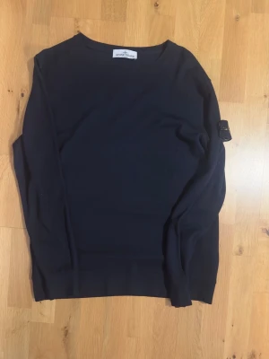 Svart tröja från Stone Island - Snygg svart tröja från Stone Island med den klassiska logotypen på ärmen. Tröjan har rund halsringning och är tillverkad i mjukt material som passar perfekt till en avslappnad stil. Enkel och stilren design som funkar till det mesta.