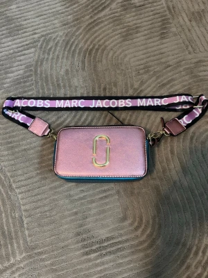 Rosa axelväska från Marc Jacobs - Cool axelväska i metallicrosa från Marc Jacobs med justerbar axelrem i svart och lila med logga. Väskan har guldiga metalldetaljer och dubbla dragkedjor upptill. Perfekt för att lyfta din outfit med en färgklick och trendig design.