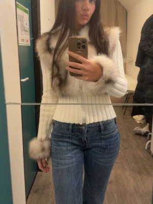 Alessa päls kofta - Säljer en vit ribbad tröja med fluffiga fuskpälsdetaljer i beige och grått runt krage och ärmslut. Tröjan har en figurnära passform och långa ärmar, perfekt för dig som vill ha en trendig och cozy look.