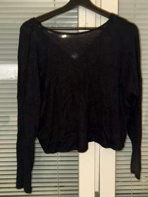 Svart långärmad tröja med spets - Svart långärmad tröja med v-ringning och dekorativ spetsdetalj längs halsringningen. Tröjan har en lös passform och är tillverkad i ett tunt, mjukt material som känns skönt mot huden. Perfekt för lager-på-lager-stil.