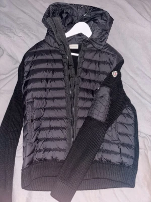 Svart cardigan från Moncler - Snygg svart cardigan från Moncler med stickade ärmar och huva. Jackan har quiltad front och rygg i glansigt material, ribbade muddar och dragkedja framtill. Moncler-logga på ärmen och praktisk ficka med dragkedja. Perfekt för kalla dagar.