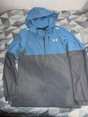 Blå och grå vindjacka från Under Armour - Snygg vindjacka från Under Armour i blått och grått med huva och hel dragkedja. Jackan har en bröstficka med dragkedja, reflekterande detaljer på axlarna och logga på bröstet. Perfekt för blåsiga dagar och sportiga outfits.