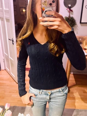 Mörkblå kabelstickad tröja Ralph Lauren - Säljer en mörkblå kabelstickad tröja från Ralph Lauren med V-ringning och lång ärm. Tröjan har en liten röd broderad logga på bröstet och ribbstickade muddar vid ärmslut och nederkant. Perfekt för en klassisk och stilren look. Kontakta för mera info!💗