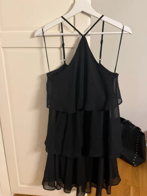 Svart volangklänning med tunna axelband - Snygg svart kortklänning med volanger och tunna, justerbara axelband som korsas framtill. Klänningen har en luftig och flowy passform, perfekt för dig som gillar en stilren och trendig look. Tillverkad i ett lätt och transparent tyg med flera lager. Inköpt i somras men aldrig använd. 