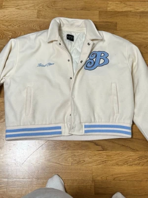 Vit bomberjacka från MAN Official - Vit bomberjacka från MAN Official med blå broderade detaljer och ribbade muddar i vitt och ljusblått. Jackan har tryckknappar framtill, broderad text på bröstet och ryggen samt en stor blå bokstav på bröstet. Perfekt för dig som vill ha en cool och sportig look.