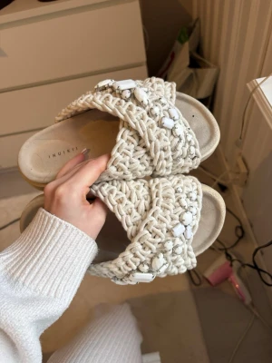 Beige flätade sandaler Inuikii - Säljer ett par beige sandaler från Inuikii med breda, flätade band och vita dekorstenar. Sandalerna har en chunky sula och öppen tå, perfekta för en trendig och avslappnad look. Materialet är textil med detaljer i plast.