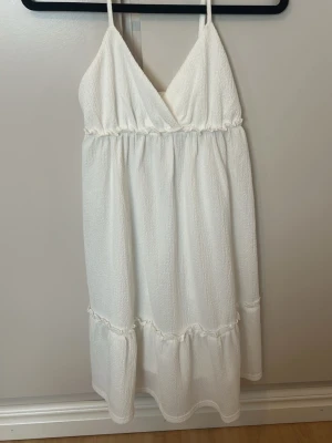 Vit omlottklänning med volang - Säljer en vit kortklänning med omlott-v-ringning och tunna axelband. Klänningen har volangdetaljer och är tillverkad i ett lätt, strukturerat material som känns som bomull. Perfekt för sommarens varma dagar.