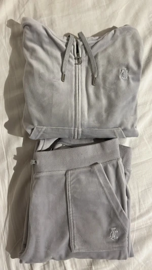 Velourset från Juicy Couture - Mysigt loungewear-set i velour från Juicy Couture. Setet består av en zip-hoodie och matchande byxor med fickor. Färgen är ljus grå med ljus lila typ? Se bild 3 för en bättre bild av färgen. Den har en liten fläck på byxorna där bak.  Antingen 699kr för båda delarna eller 450kr styck. 