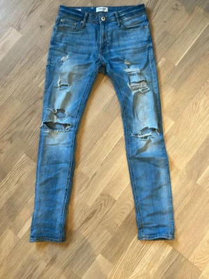 Blå slitna jeans från Jack & Jones  - Säljer ett par blå jeans med rejält slitna och trasiga detaljer framtill. Klassisk femficksmodell med låg midja och smal passform. Perfekta för en avslappnad och trendig streetstil.