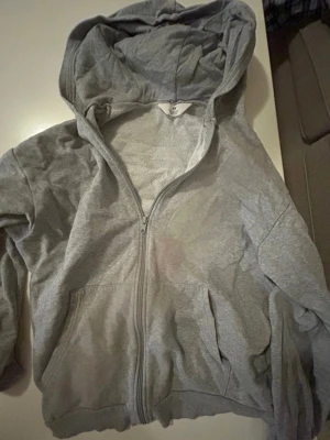 Grå zip hoodie med huva - H&M - En klassisk grå hoodie med dragkedja framtill och stor huva. Hoodien har två fickor på magen och är tillverkad i mjukt sweatshirtmaterial. Perfekt för en avslappnad och chill stil.