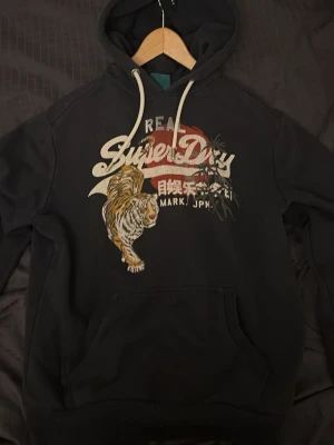 Mörkblå Superdry hoodie med unik design  - Mörkblå hoodie från Superdry med stort tryck framtill, där loggan kombineras med en cool tiger och japanska tecken. Klassisk känguruficka och tjocka vita nören i huvan. Perfekt för dig som vill ha en snygg och stilren hoodie samtidigt som den är unik!