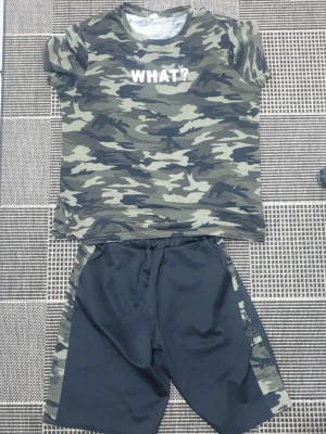 Kamouflage set med t-shirt och shorts - Säljer ett coolt set med t-shirt och shorts. T-shirten har grönt kamouflagemönster och texten 'WHAT?' framtill. Shortsen är svarta med kamouflagedetaljer på sidorna. Perfekt för en avslappnad och sportig stil.
