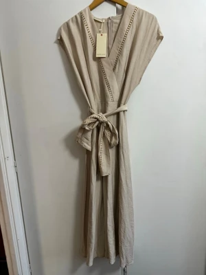 Beige omlottklänning med bälte  - Snygg beige långklänning från Madam Rouge med omlottmodell och knytbälte i midjan. Klänningen har v-ringning med dekorativa detaljer längs kanten och är ärmlös. Perfekt för en avslappnad och trendig look.