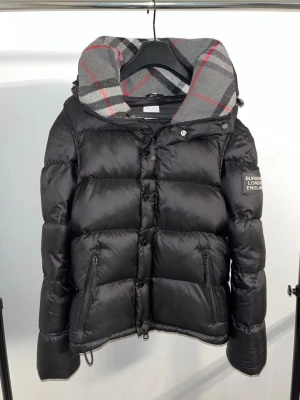 Svart dunjacka från Burberry - Snygg svart dunjacka från Burberry med klassisk rutmönstrad huva i grått, rött och svart. Jackan har dragkedja och knappar framtill, två fickor med dragkedja och Burberry-logga på ärmen. Perfekt för kalla vinterdagar och kan göras till en väst genom att dra ner kedjorna som finns på insidan av ärmen!