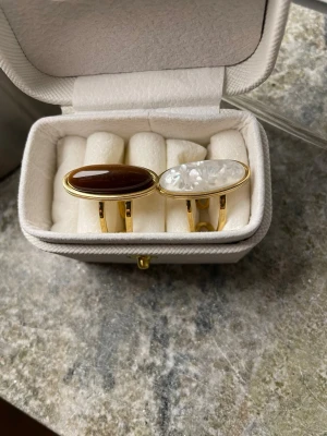 Guldfärgade och rostfria statementringar med stenar - Två coola ringar i Copper rostfria, en med avlång brun sten och en med avlång vit pärlemorliknande sten. Båda har modern, minimalistisk design och passar perfekt för att lyfta din stil. Både har öppning och kan justeras i storleken. Måttet 1,8 cm diameter 