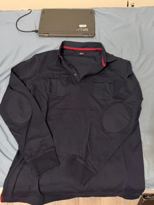 GEREM collection mörkblå långärmad pikétröja - Snygg mörkblå långärmad pikétröja från Jerseem med krage, knappar och två bröstfickor. Tröjan har ribbade muddar vid ärmsluten och förstärkta armbågar. Röd detalj på insidan av kragen ger en extra touch. Perfekt för en avslappnad men stilren look.