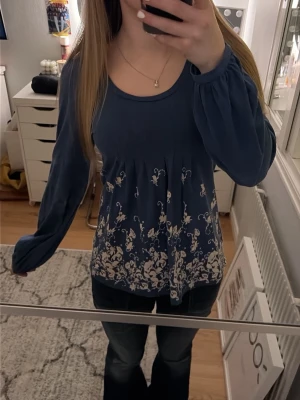 Blå blus med vita blommor - Söt blå blus med långa ärmar och vita blommönster nertill. Blusen har rund halsringning och ett lätt utställt fall som ger en avslappnad look. Perfekt att matcha med jeans för en trendig vardagsstil.