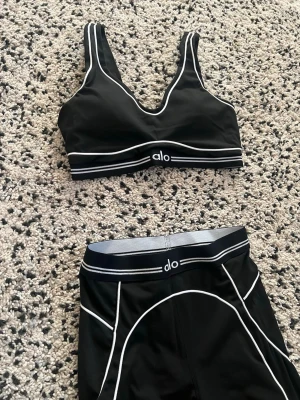 Svart träningsset från Alo - Snyggt svart träningsset från Alo med sport-bh och höga tights. Setet har vita kontrastsömmar och logga på både bh och midja. Materialet är stretchigt och passar perfekt för gym, yoga eller löpning. Sportig och modern look.