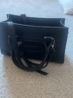 Svart handväska i konstläder - Snygg svart handväska i konstläder med dubbla handtag och avtagbar axelrem. Väskan har en liten ytterficka med dragkedja och silverfärgade detaljer. Stilren och enkel design som passar till många outfits.