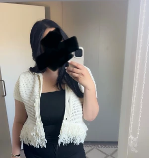 Vit stickad kort kofta med fransar - Säljer en vit stickad kort kofta med knappar framtill och dekorativa fransar längs nederkanten. Koftan har korta ärmar och en öppen, luftig stickning som ger en trendig look. Perfekt att slänga över ett linne eller t-shirt.