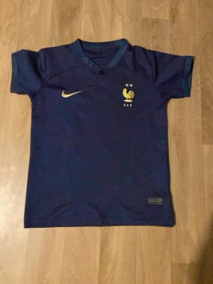 Mbappe fotbollströja Frankrike  - Mörkblå officiell Frankrike fotbollströja från Nike med guldfärgade detaljer, FFF-logga och två stjärnor på bröstet. På ryggen står det Mbappé och nummer 10 i guld. Perfekt för dig som älskar fotboll och vill stötta landslaget.